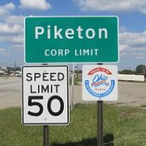 Green road sign saying Piketon Corp Limit, another sign saying Speed Limit 50 and a sign with an Ohio logo on it and in the background a highway and some grass and the blue sky with white clouds
