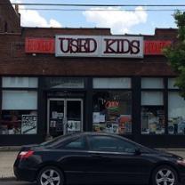 Brick building with sign at top saying Used Kids and a storefront with windows, a black car parked in front