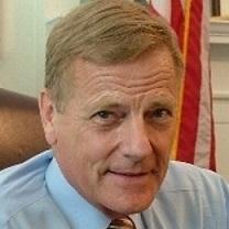 Close up of white man's face with brown hair and blue button down shirt and tie in front of a flag, his eyebrows a bit raised