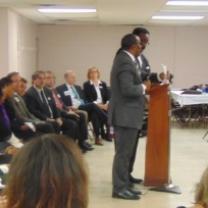 Lots of men in suits sitting in a row on chairs facing right and two black men in suits standing at a podium