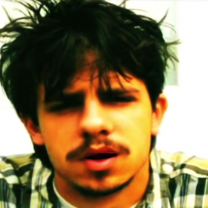 Close up of a young white man's face with dark hair askew and a moustache and goatee