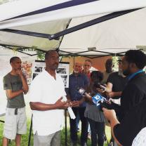 A press conference with a black man in a suit holding a mic for another black man with glasses and a white shirt to talk into and other people standing around watching