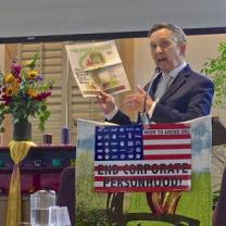 Dennis Kucinich, 60-ish white guy in a suit at a podium holding up a copy of the Free Press