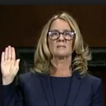 Blonde woman with glasses holding her hand up as if swearing to tell the truth